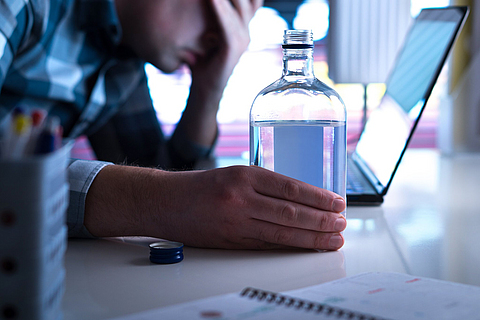Ein Mann sitzt mit Alkoholflasche vor dem Laptop und sieht bedrückt aus.