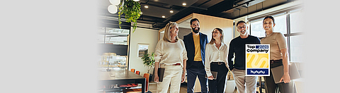 Fünf Kollegen stehen beieinander in einem Büro und lachen. Im Vordergrund das Kununu Top Company Logo.