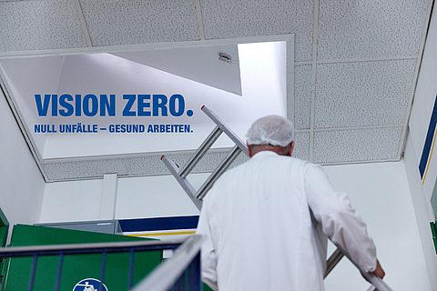 Ein Mann stellt eine Leiter an eine Luke an. Daneben steht "Vision Zero. Null Unfälle - gesund arbeiten."